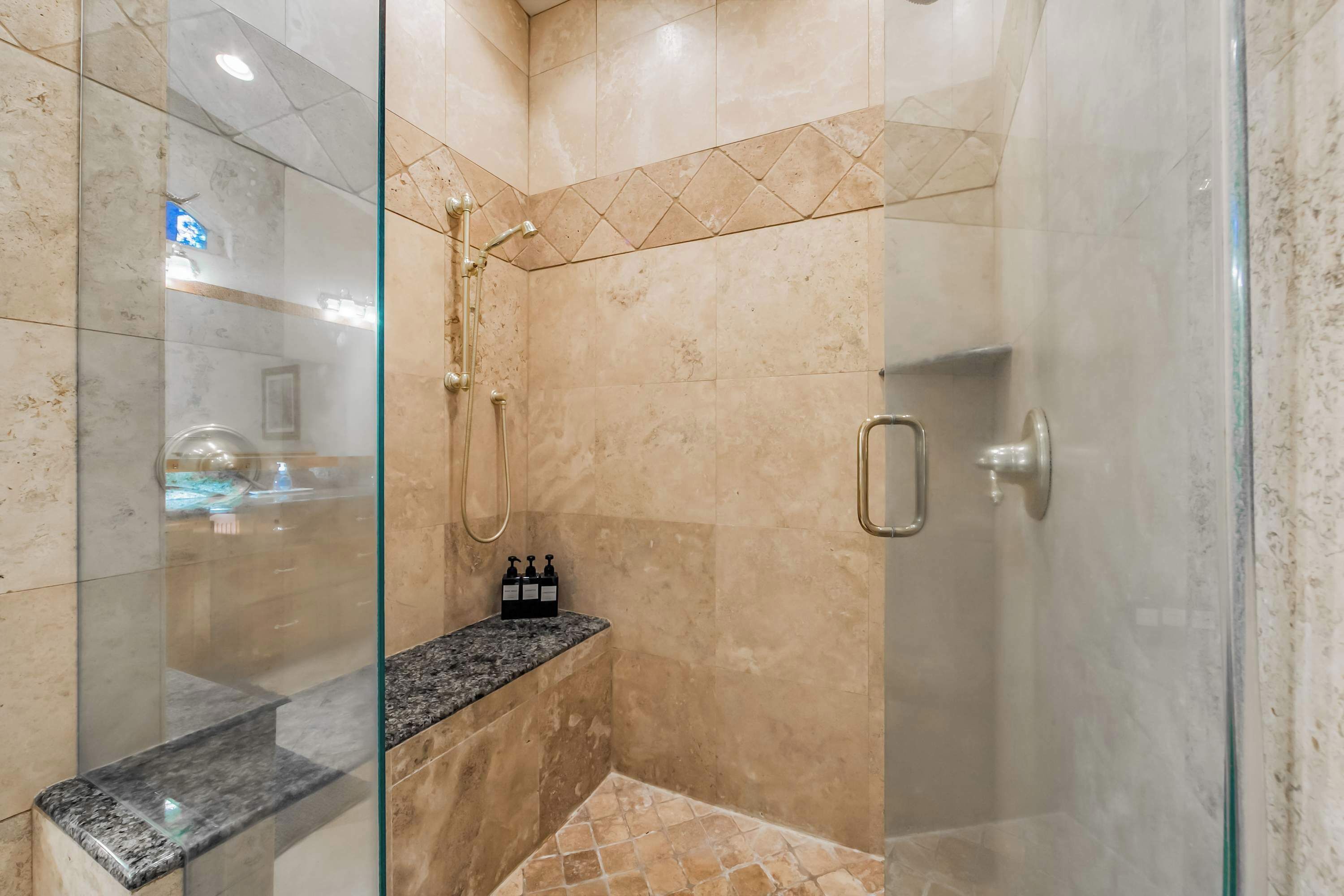Une douche fermée par des parois de verre, avec un mur en marbre, un banc et une bouteille de shampoing.