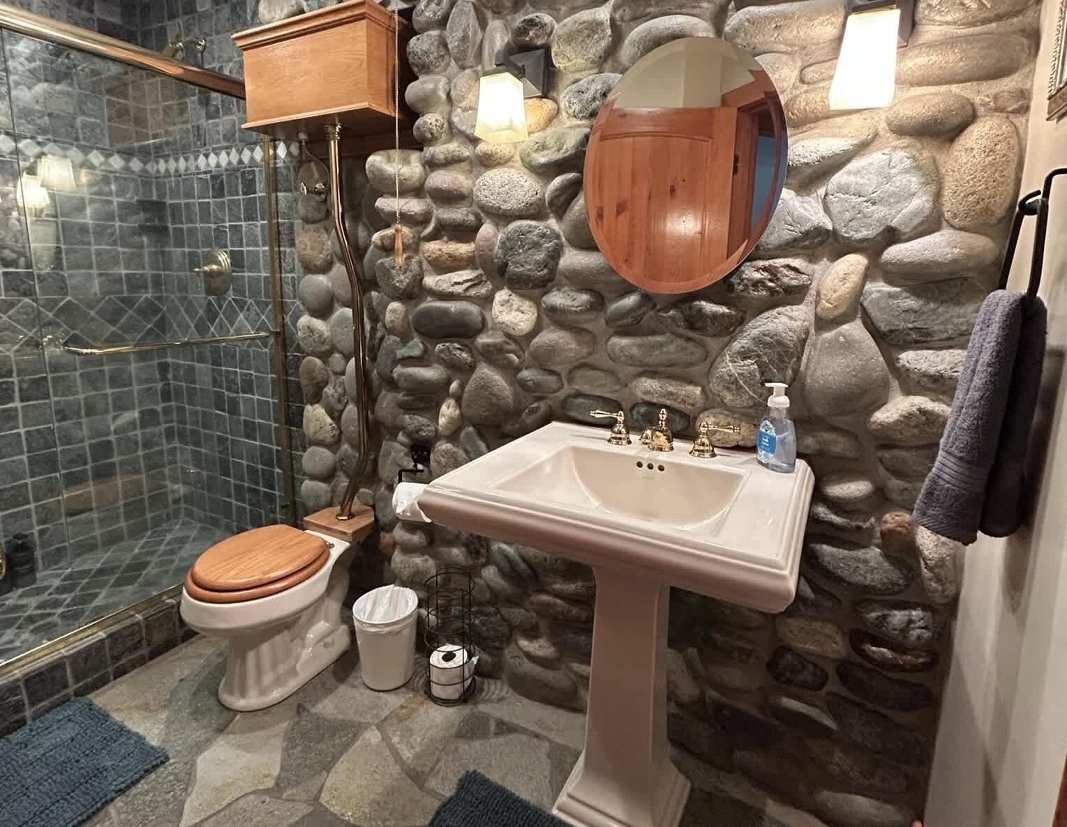 Un baño con pared de piedra, lavamanos blanco, inodoro de madera y cabina de ducha de vidrio.