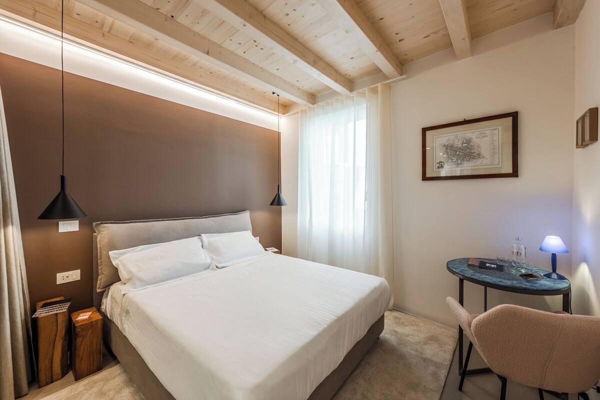 A neatly arranged bedroom with a bed, bedside lamps, a small table with a lamp, and a framed picture on the wall.