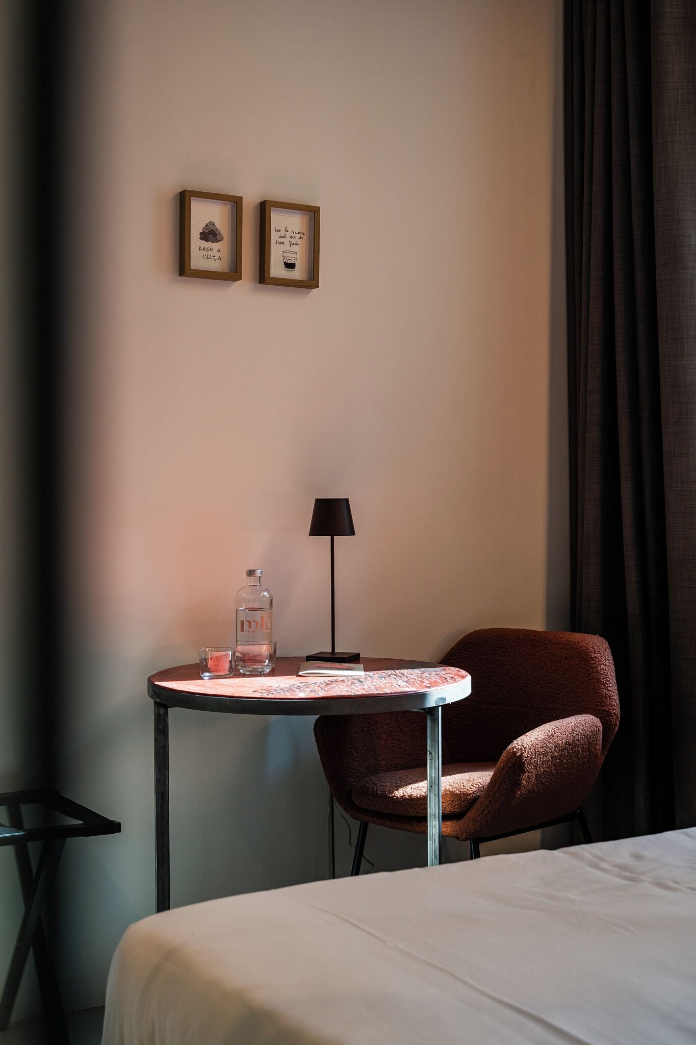 A hotel room with a bed, a small table with a lamp, a chair, and two framed pictures on the wall.