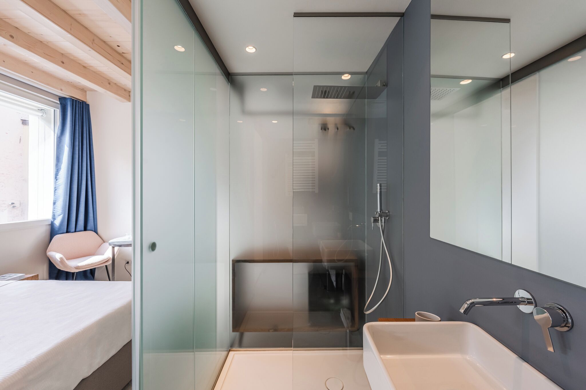 A modern bathroom with a glass shower enclosure, a white sink, and a bed with a chair.