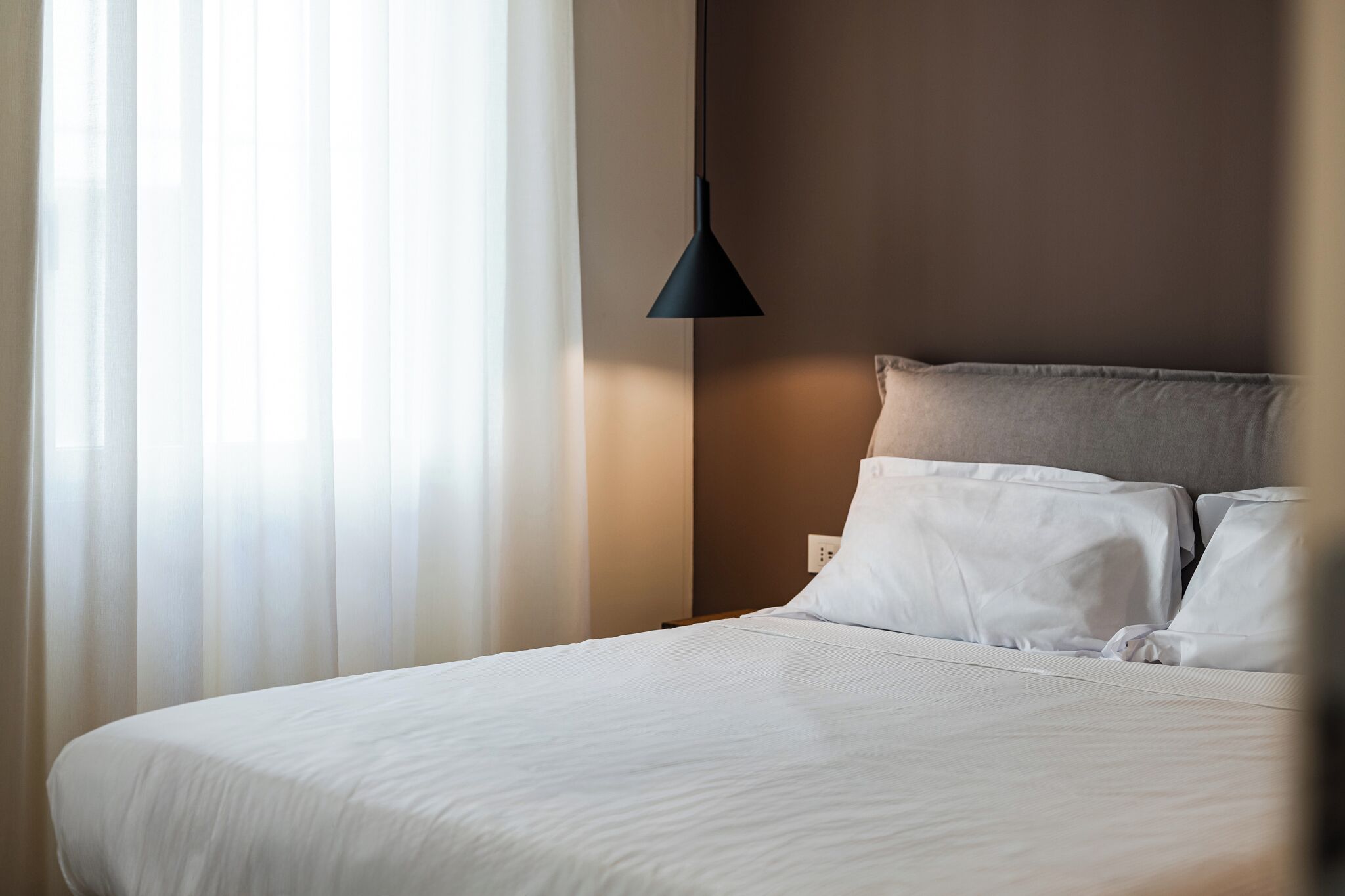 Una cama bien hecha con sábanas blancas, una lámpara colgante oscura y cortinas translúcidas que dejan entrar luz natural.