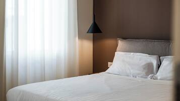 Una cama bien hecha con sábanas blancas, una lámpara colgante oscura y cortinas translúcidas que dejan entrar luz natural.