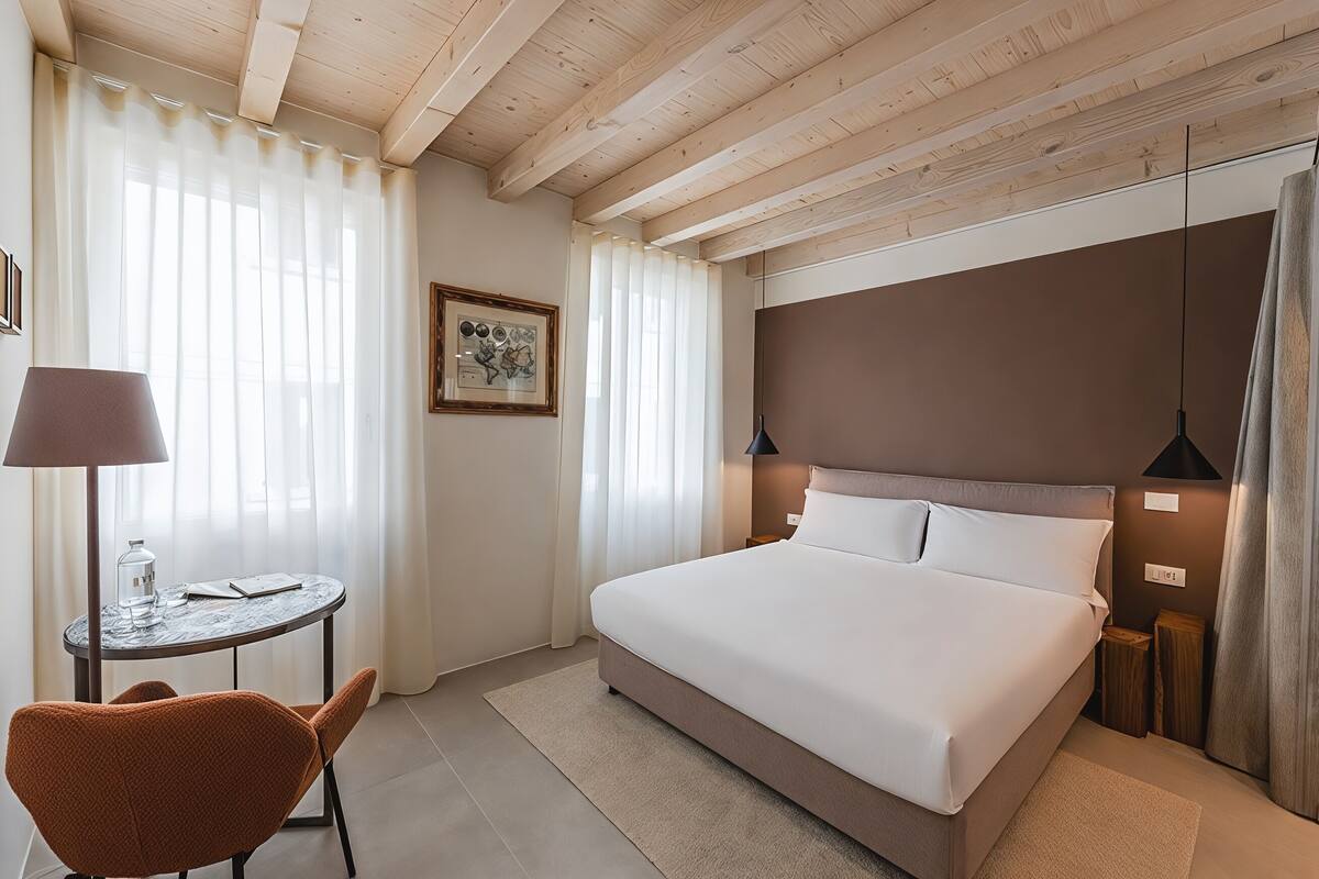 A hotel room with a large bed, a round table with a lamp, a chair, and a framed picture on the wall.
