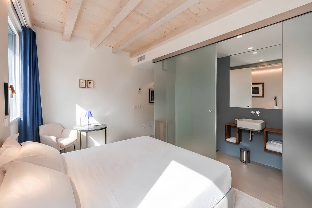 A modern hotel room with a large bed, a desk, a chair, a bathroom with a mirror, and a sink.