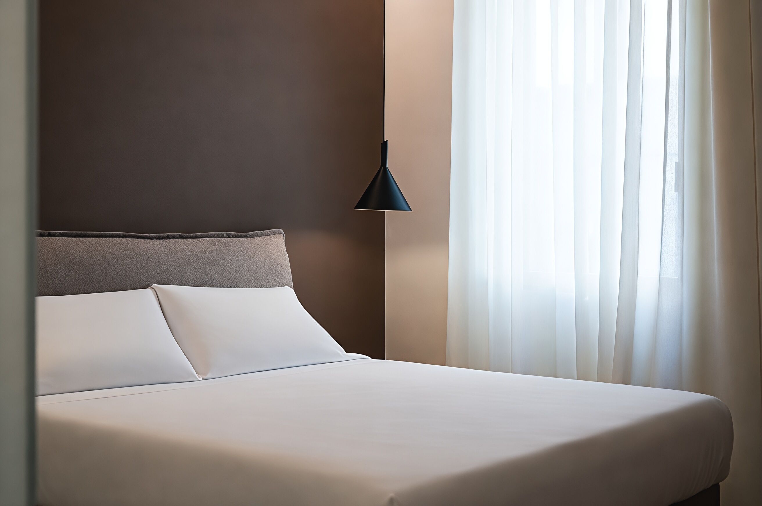 A hotel room with a bed, a pillow, a headboard, a wall, and a window with curtains.
