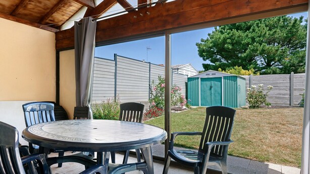 A covered patio with a round table and chairs, a sliding glass door, and a view of a backyard with a shed and a tree. - Family House with 4 Bedrooms for 13 Guests (La Plaine-sur-Mer)