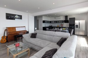 Ein modernes Wohnzimmer mit einem grauen Sofa, einem Glastisch, einem Holzpiano und einer Küche im Hintergrund.