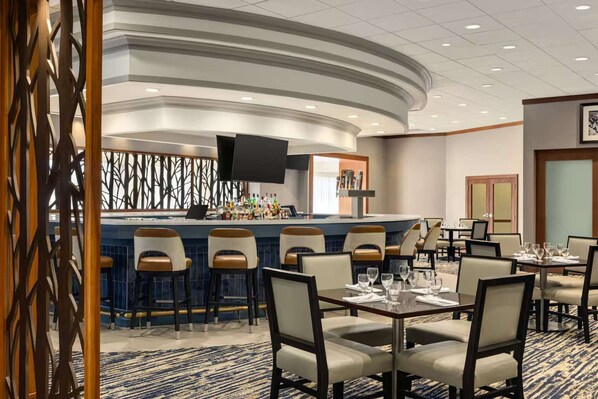 A hotel bar with a curved counter, bar stools, and a television screen. There are dining tables with chairs arranged in the room.