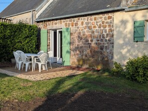 Un bâtiment en pierre avec une porte et des fenêtres, une table avec des chaises et une haie.