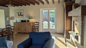 A living room with a blue sofa, wooden beams, a fireplace, and a dining area with a table and chairs. - Family home 500 m from the beach (Saint-Marcouf)