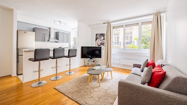 A modern living room with a grey sofa, a TV, and a kitchen area with bar stools. - Mobility Lease - Porte Dauphine - 1 large bedroom (Paris)