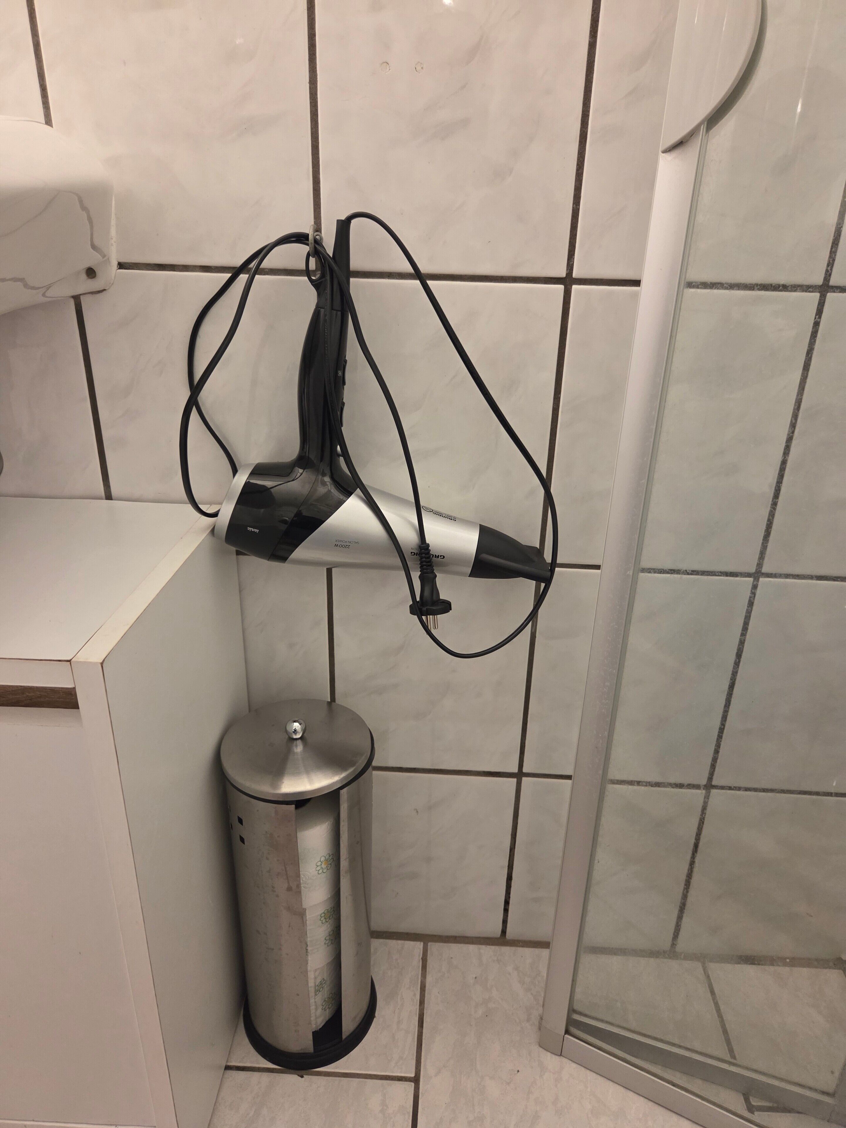 A bathroom with a hairdryer mounted on the wall, a trash bin, and tiled walls.
