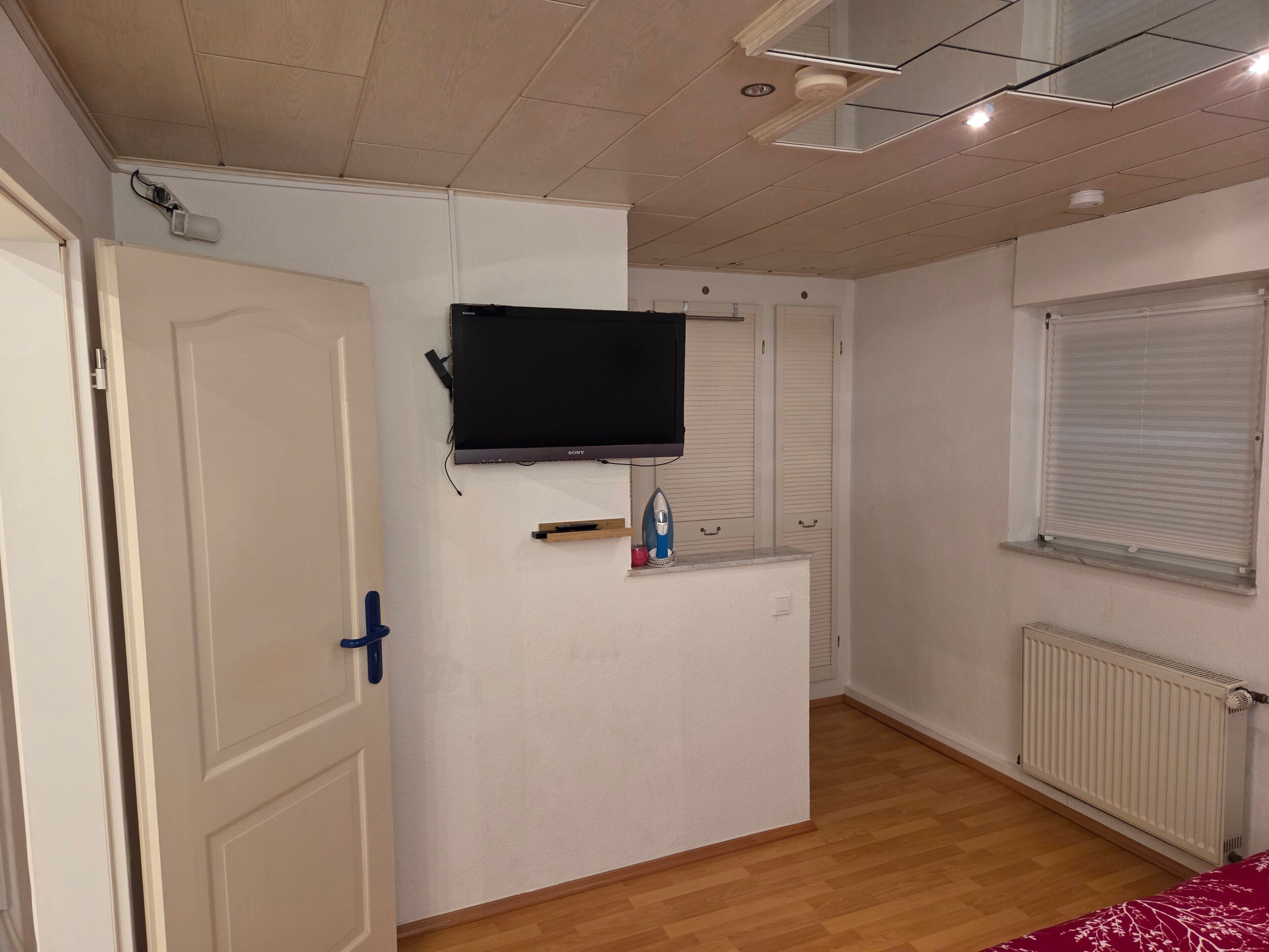A room with a flat-screen TV mounted on the wall, a radiator, and a window with blinds.