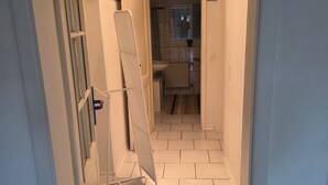 Un couloir étroit au sol carrelé, une porte blanche sur la gauche et une fenêtre au fond.