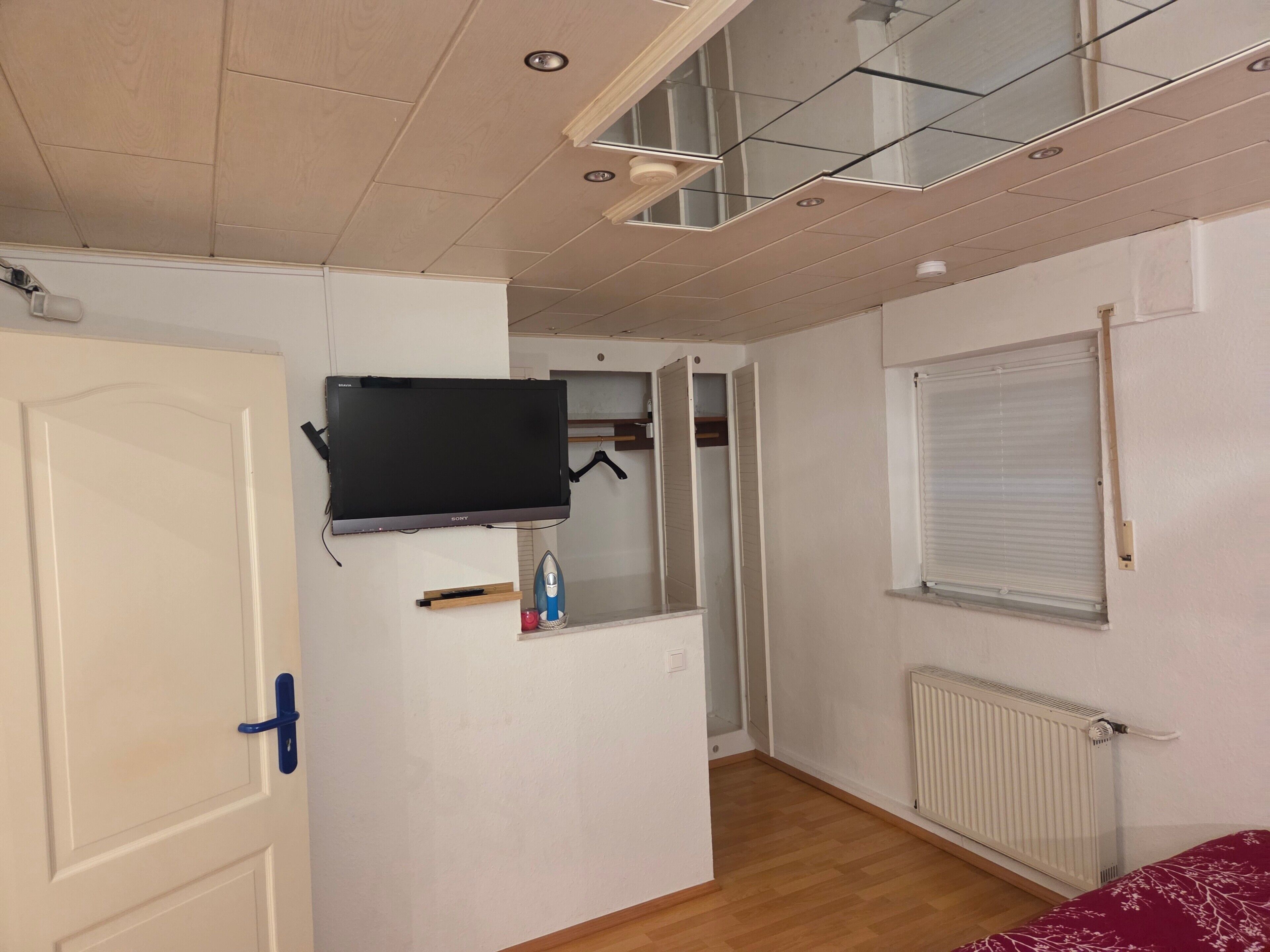A room with a flat-screen TV, a wardrobe, a radiator, and a window with blinds.