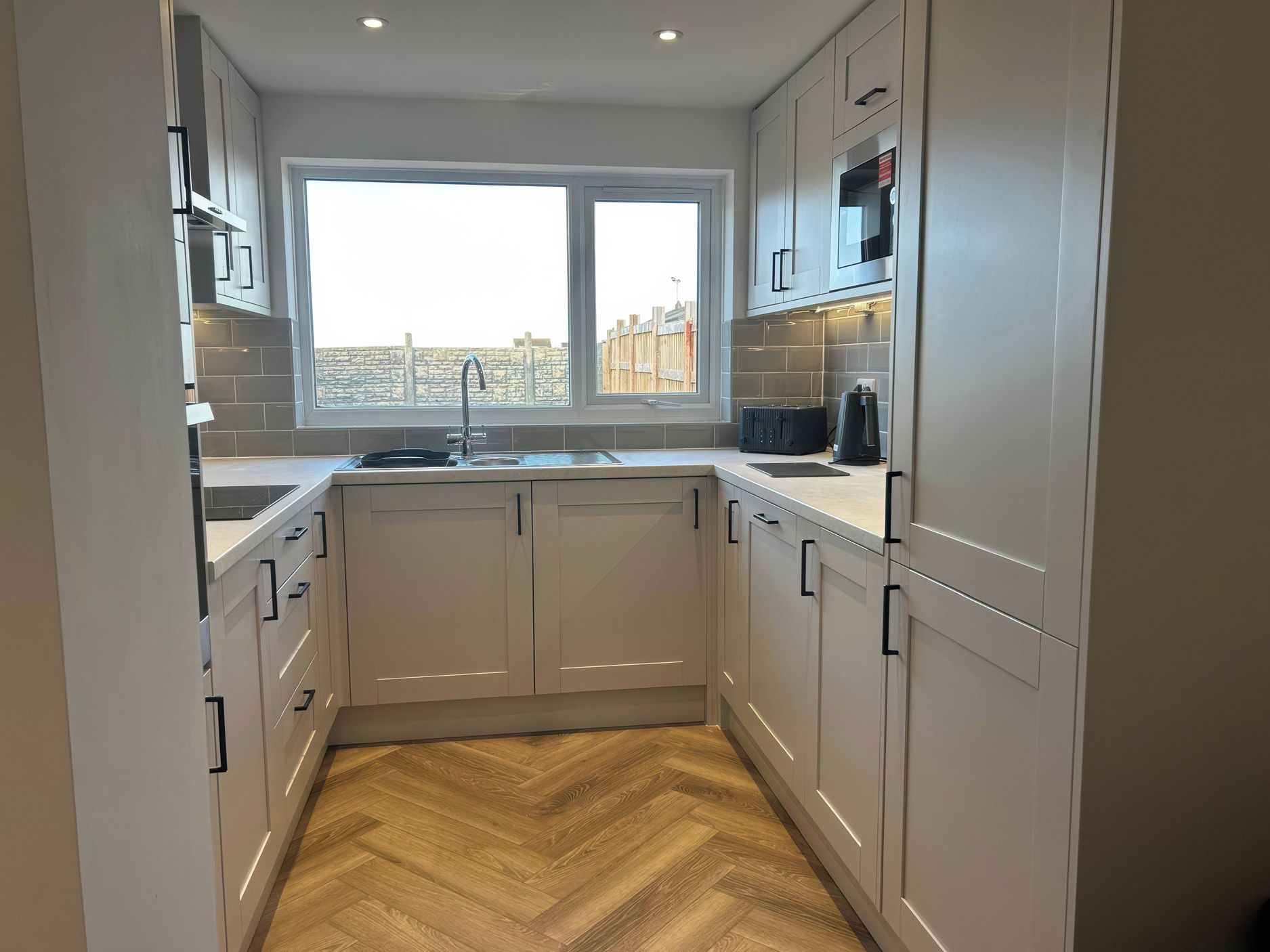 A modern kitchen with white cabinets, a herringbone wood floor, a window with a view, and a built-in microwave.