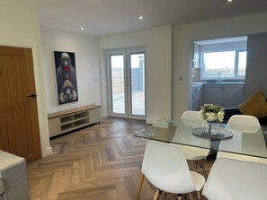 Un comedor moderno con una mesa de vidrio y sillas blancas, piso de madera, una cocina con ventana y una puerta que da a un balcón.