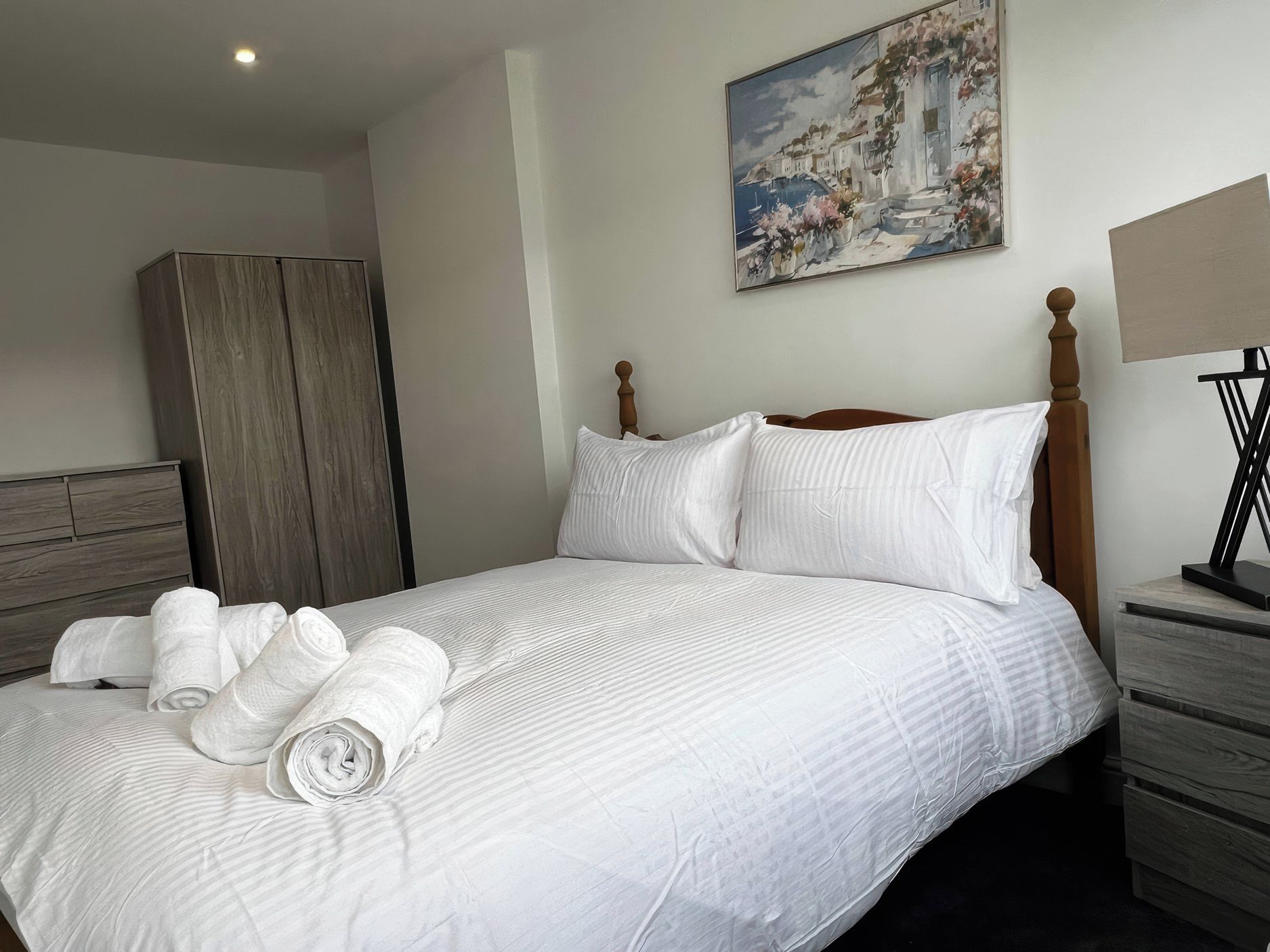 Una cama bien hecha con ropa blanca y toallas enrolladas, un cabecero de madera, un cuadro en la pared y una mesita de noche con una lámpara.