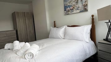 Una cama bien hecha con ropa blanca y toallas enrolladas, un cabecero de madera, un cuadro en la pared y una mesita de noche con una lámpara.