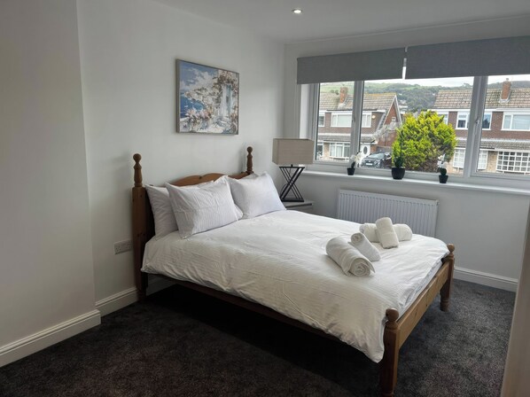 Una cama bien hecha con ropa de cama blanca y toallas enrolladas, un cuadro en la pared, una ventana con vistas a casas y un radiador.