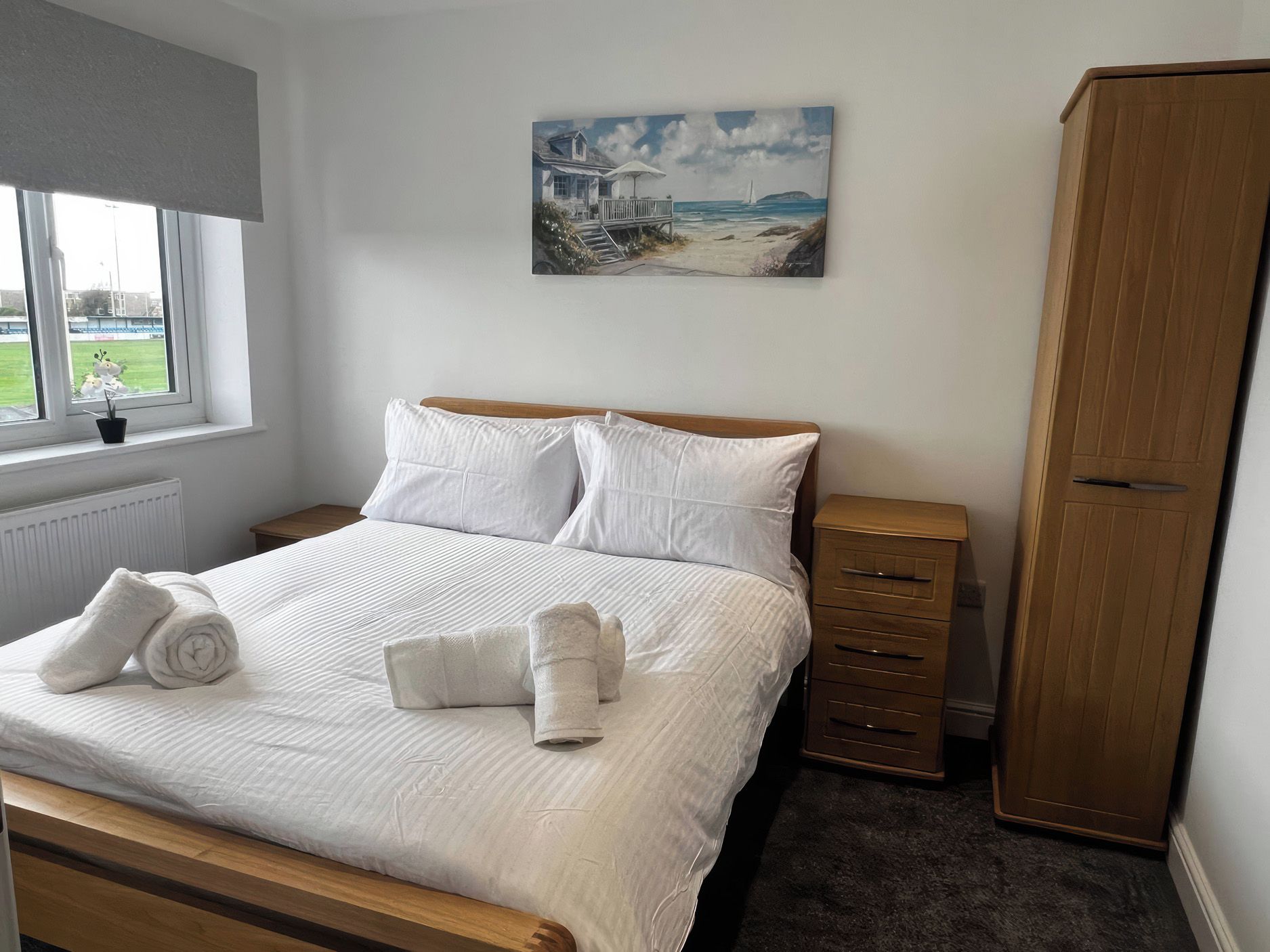 A bedroom with a bed, a nightstand, a wardrobe, a window with a view of a grassy field, and a painting on the wall.