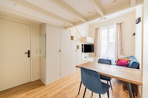 Ein kleines Zimmer mit einem Holztisch, zwei Stühlen, einem an der Wand montierten Fernseher und einem Fenster mit Vorhängen.