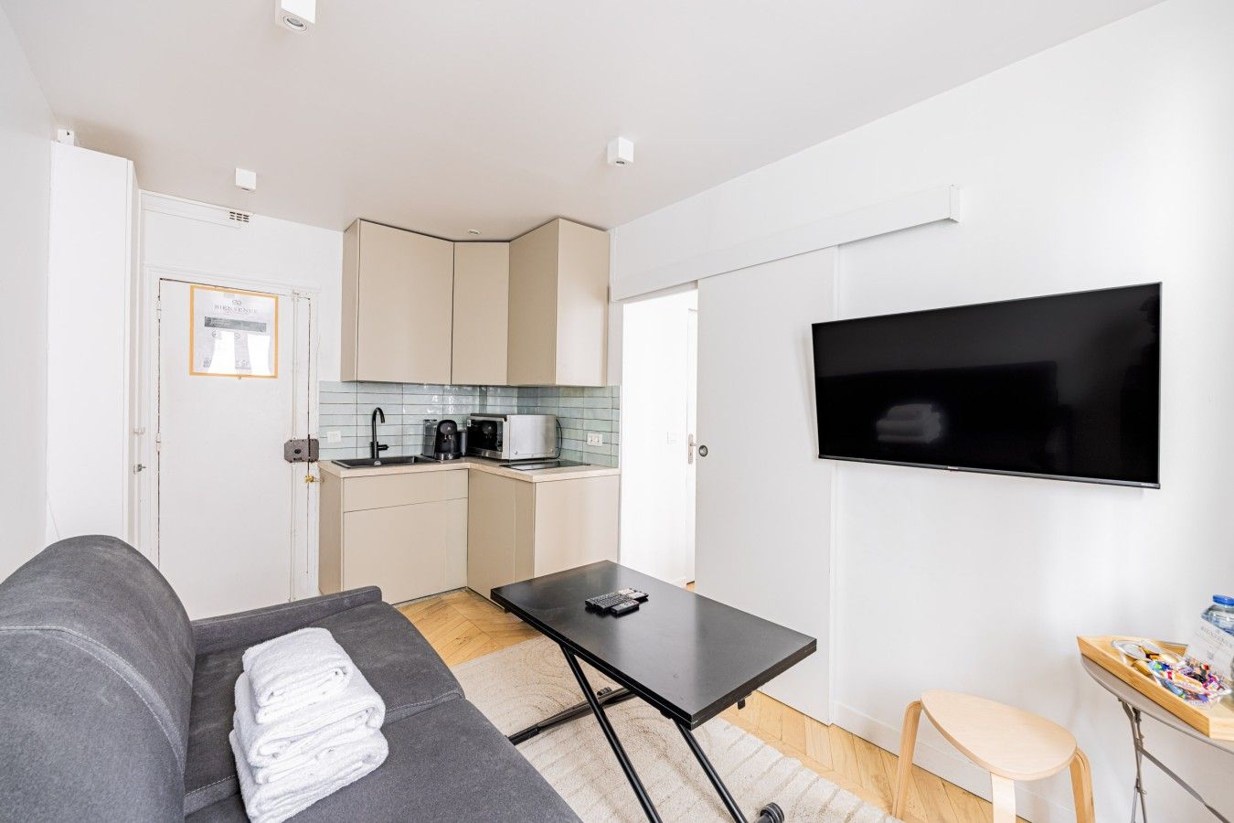 A compact living space with a kitchenette, a flat-screen TV, a sofa, a small table, and a chair.