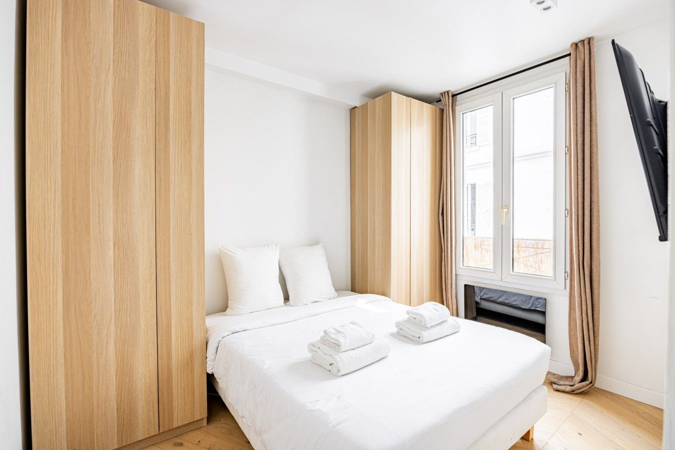Ein Schlafzimmer mit einem Bett, weißer Bettwäsche, einem Holzschrank, einem Fenster mit Vorhängen und einem Flachbildfernseher.