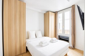 Ein Schlafzimmer mit einem Bett, weißer Bettwäsche, einem Holzschrank, einem Fenster mit Vorhängen und einem Flachbildfernseher.
