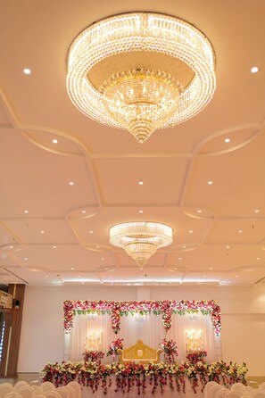 Un salón amplio con candelabros, un escenario con adornos florales y filas de sillas blancas.