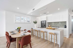 A modern kitchen with white cabinetry, a central island with stools, and a dining area with a table set for four. - Kaze South in Vincentia (Vincentia)