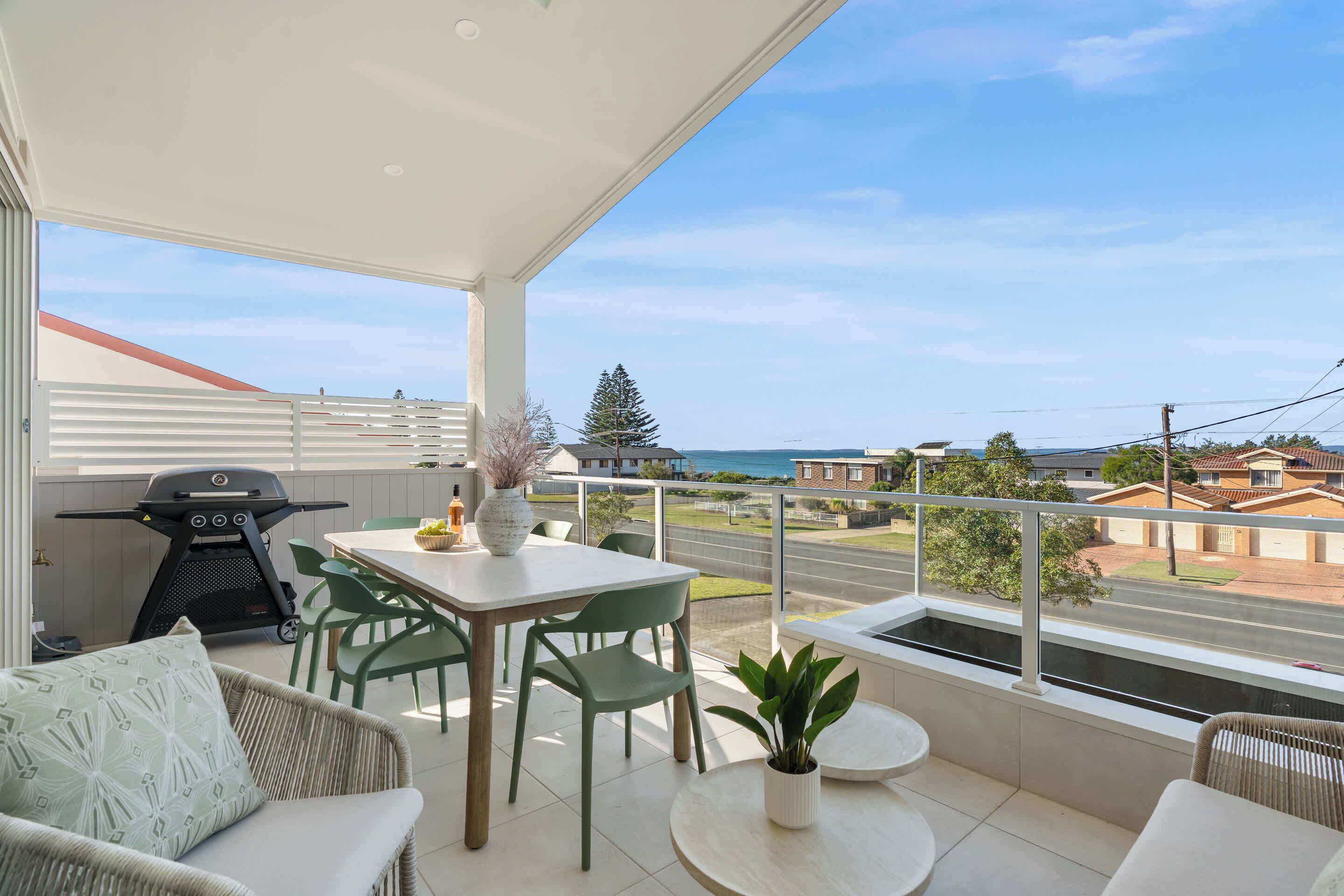 A balcony with a table, chairs, a grill, and a view of the ocean and neighborhood.