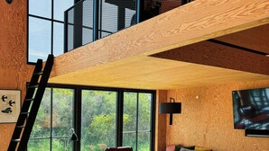 A loft-style living space with a wooden interior, large windows, a mezzanine level, and a television mounted on the wall. - Creative summer house 200 metres from beach (Sjællands Odde)