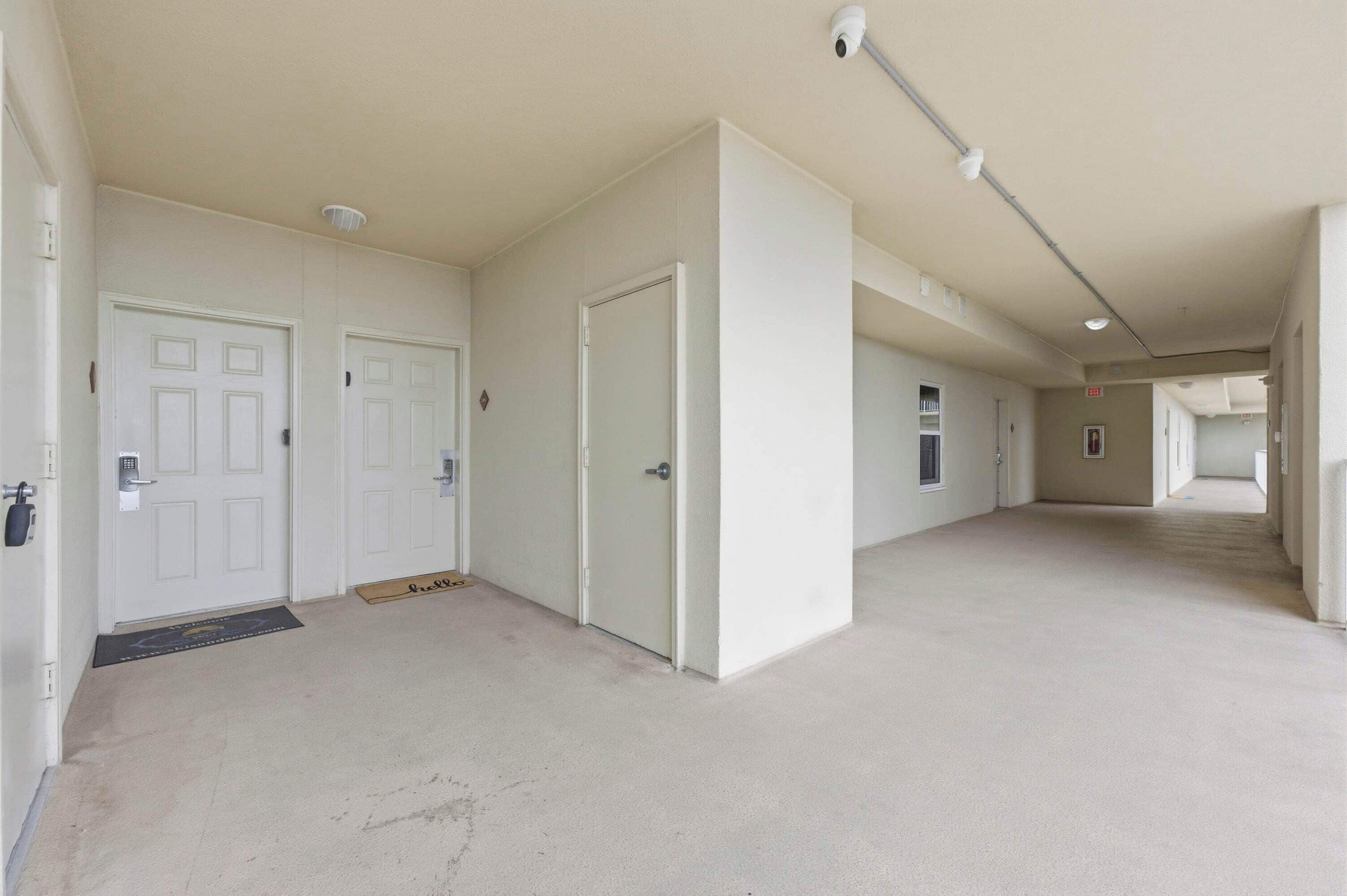Un couloir avec des portes blanches, une caméra de surveillance et un paillasson.