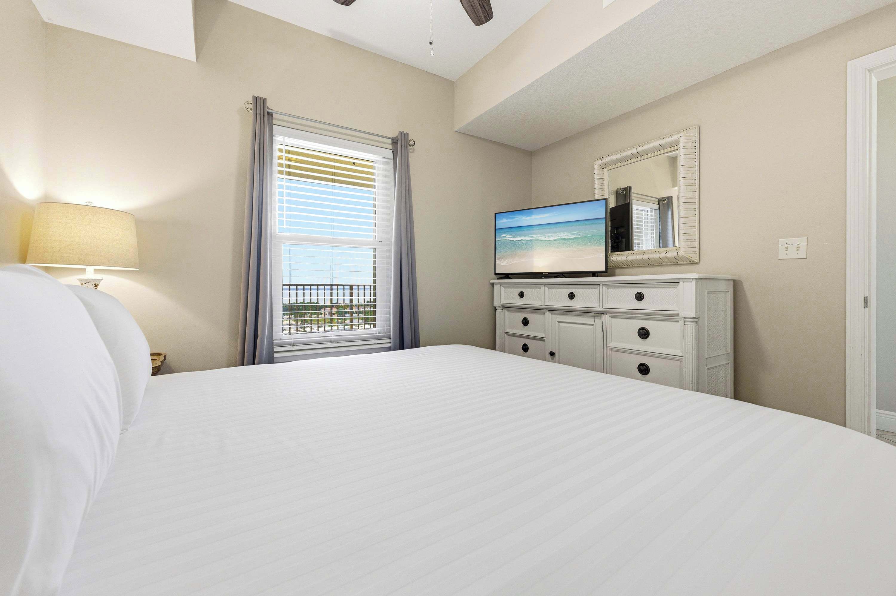 Habitación de hotel con cama, televisor en una cómoda, ventana con persianas y vista al balcón.