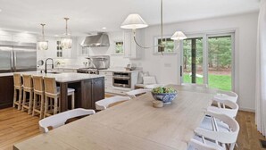A modern kitchen with a large island, bar stools, a dining table with chairs, and a view of the outdoors.