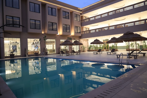 Una piscina con sillas de descanso y sombrillas, rodeada por un edificio de varios pisos con balcones.