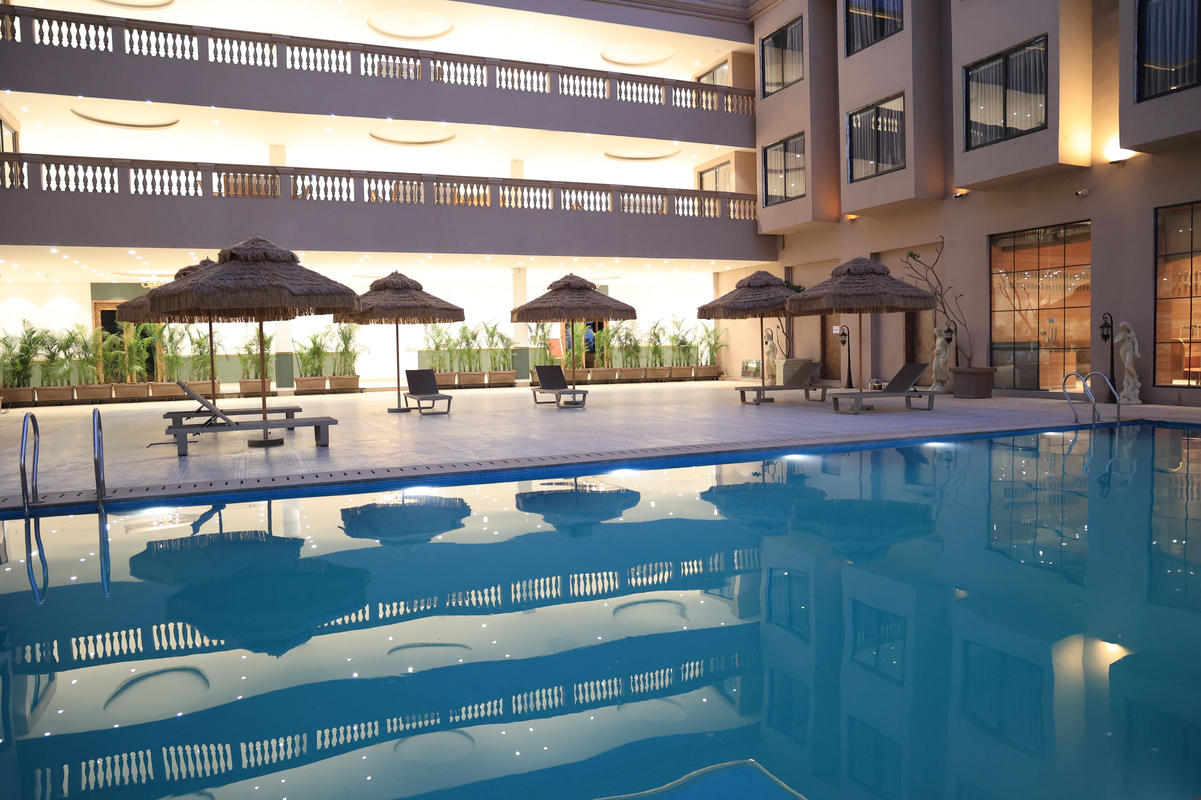 Piscina com espreguiçadeiras e guarda-sóis de palha, rodeada por um edifício com vários níveis e varandas.