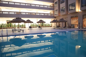 A swimming pool with lounge chairs and thatched umbrellas, surrounded by a multi-level building with balconies.