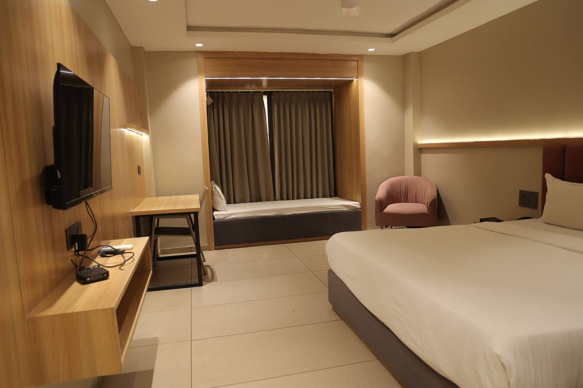 A hotel room with a bed, a desk, a chair, a TV mounted on the wall, and a window with curtains.