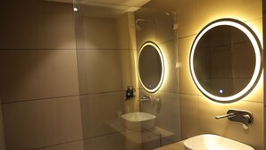 Une salle de bain moderne avec une douche à parois de verre, un grand miroir rond doté d’un éclairage indirect et un lavabo blanc.