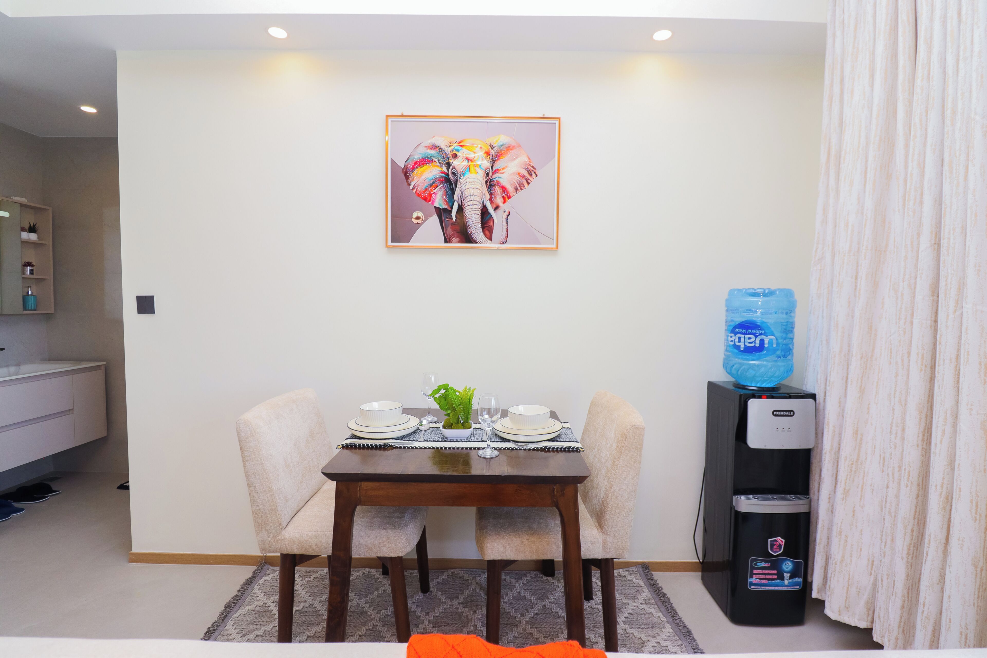 Un comedor con una mesa preparada para dos, un dispensador de agua y una obra de arte enmarcada en la pared.