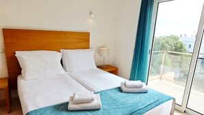 Una cama doble con sábanas blancas, un plaid azul y dos toallas dobladas al lado.