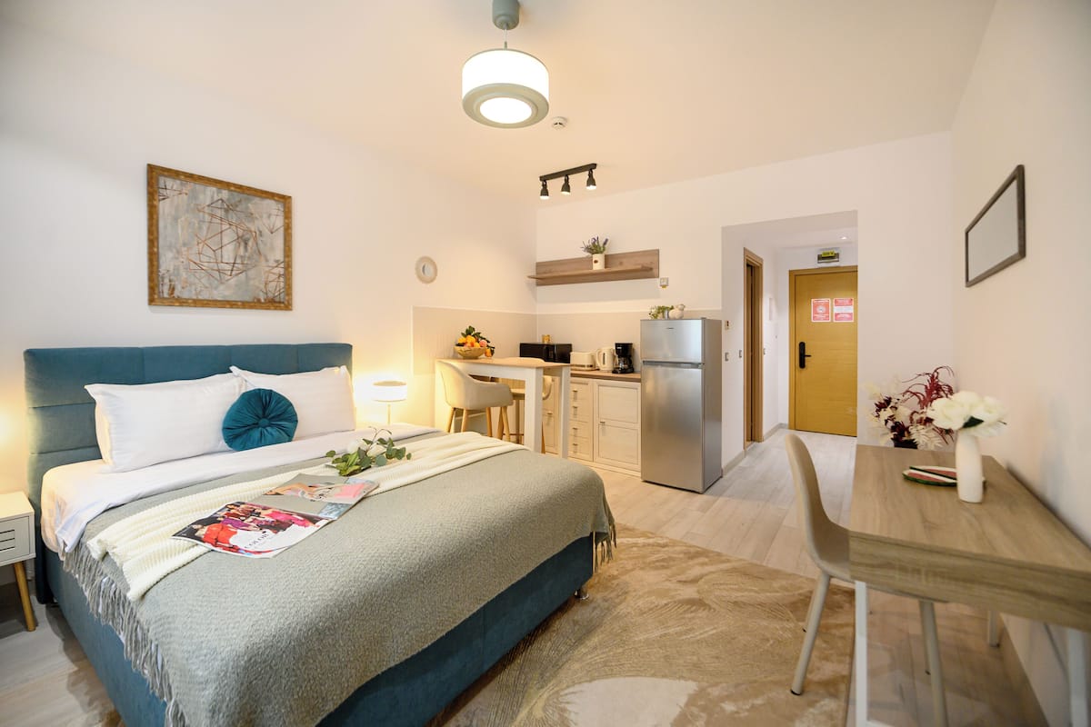 A modern hotel room with a bed, bedside table, desk, and kitchenette.