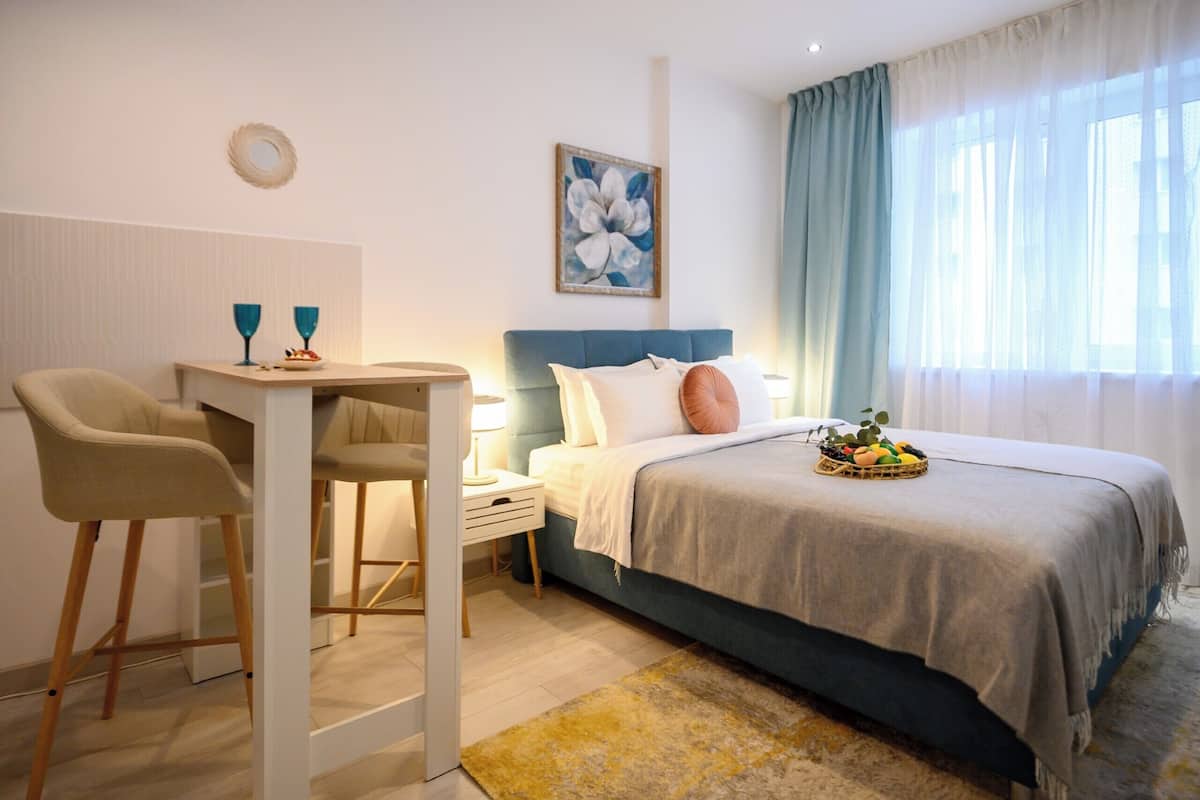 A bedroom with a bed, a desk, two chairs, a nightstand, a wall art piece, and a window with curtains.