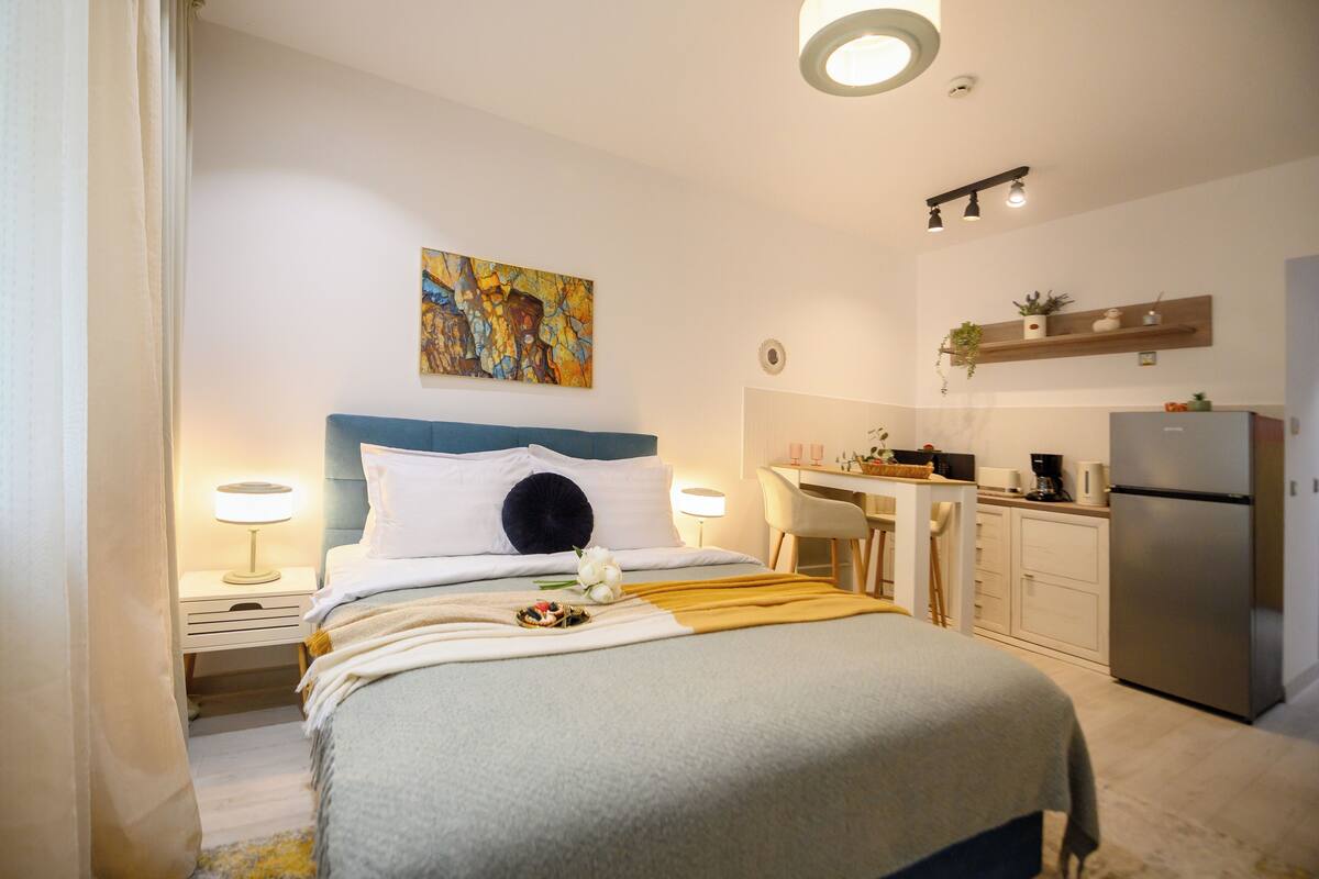A hotel room with a bed, bedside lamps, a desk, a chair, a kitchen area, and a painting on the wall.
