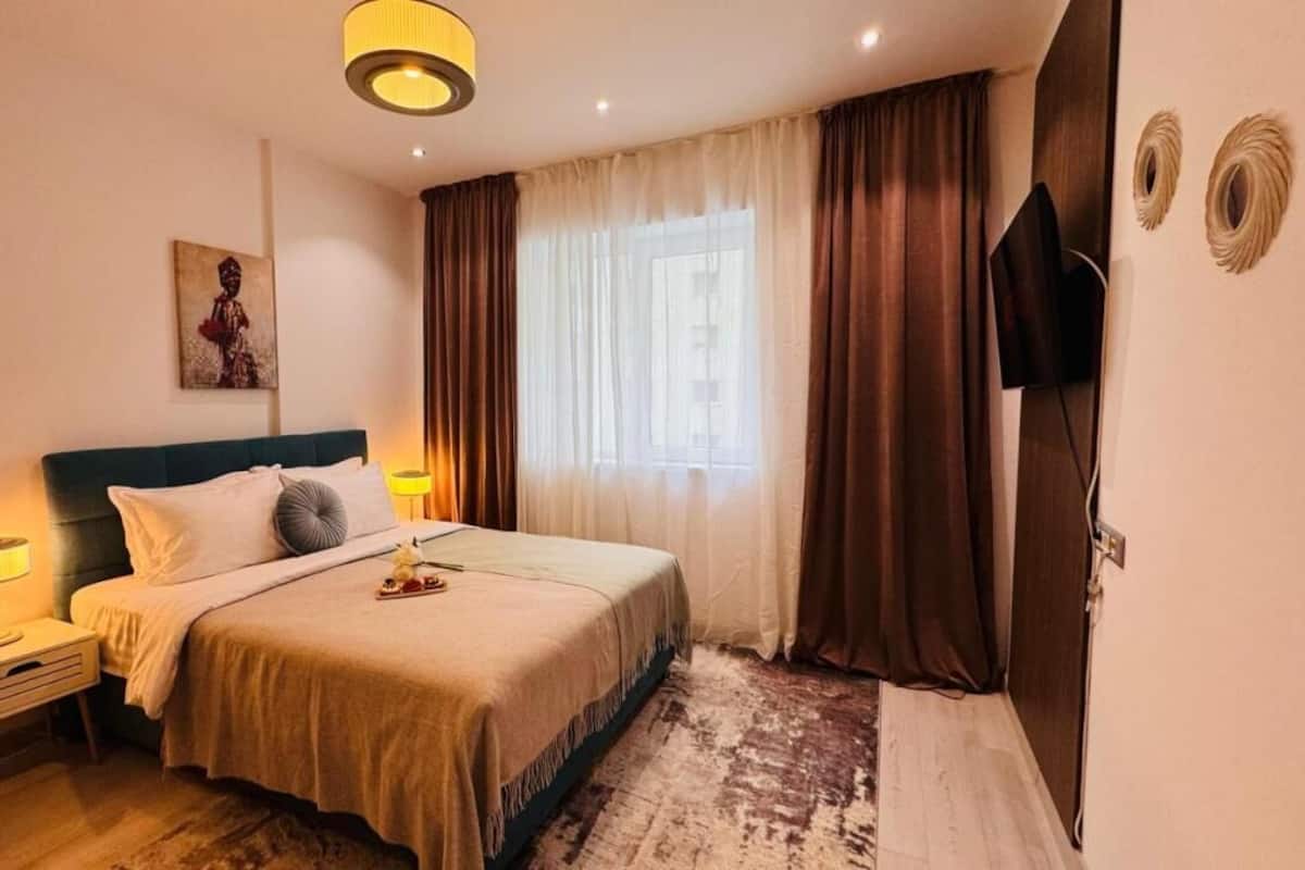 A hotel room with a bed, bedside lamps, a TV, a painting on the wall, and a window with curtains.