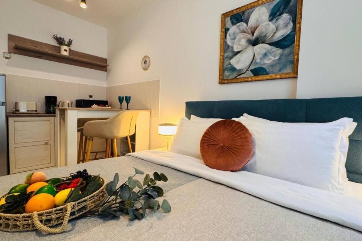 A bedroom with a bed, a bedside table, a fruit basket, a chair, a desk, a kitchen area, and a painting on the wall.