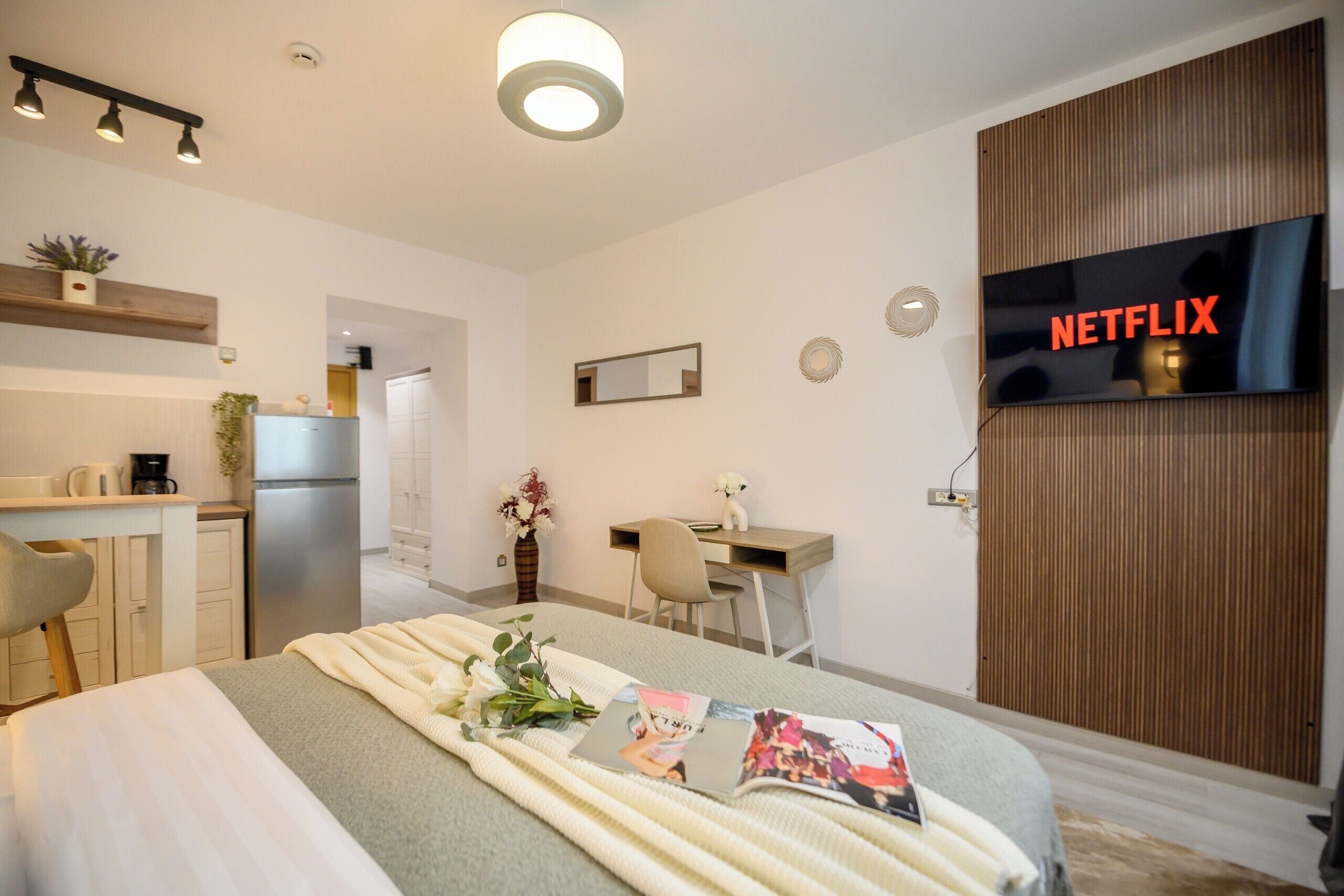 A modern hotel room with a bed, a desk, a kitchenette, and a TV displaying 'NETFLIX'.